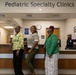 Three women are posing together at the reception desk of a Pediatric Specialty Clinics facility. They appear to be staff members or healthcare professionals, dressed in professional attire and smiling for the photo. The setting is a well-lit clinical reception area with standard medical office furnishings.
