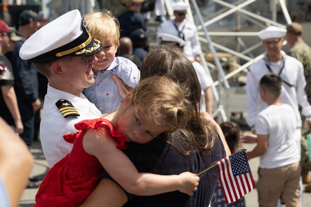 A U.S. Navy officer in dress white uniform is reuniting with his family at what appears to be a homecoming ceremony at a naval base. He is holding a young boy while a woman embraces a young girl in a red dress who is holding a small American flag. The background shows other sailors in uniform and civilians gathered at the pier.