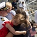 A U.S. Navy officer in dress white uniform is reuniting with his family at what appears to be a homecoming ceremony at a naval base. He is holding a young boy while a woman embraces a young girl in a red dress who is holding a small American flag. The background shows other sailors in uniform and civilians gathered at the pier.