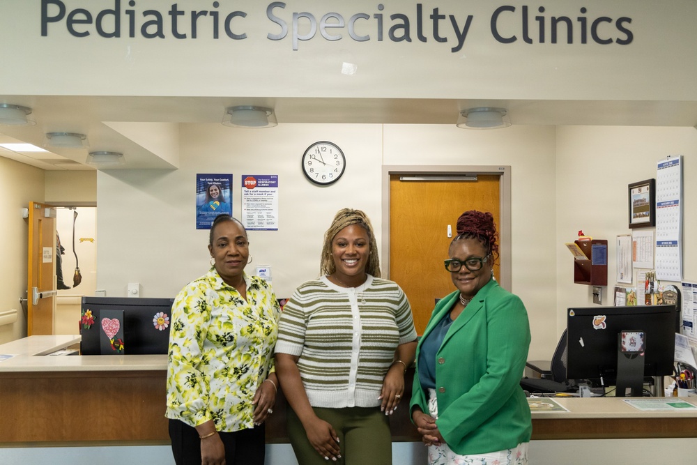 Three women are posing together at the reception desk of a Pediatric Specialty Clinics facility. They appear to be staff members smiling for a professional or commemorative photo. The setting is a well-lit medical clinic reception area with signage, a clock, and administrative equipment visible.
