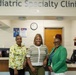 Three women are posing together at the reception desk of a Pediatric Specialty Clinics facility. They appear to be staff members smiling for a professional or commemorative photo. The setting is a well-lit medical clinic reception area with signage, a clock, and administrative equipment visible.