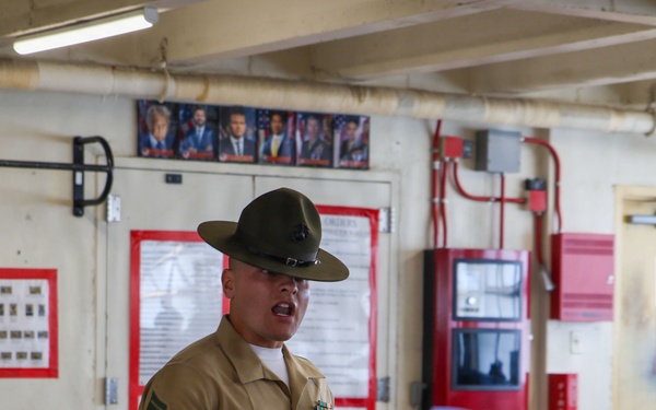 A U.S. Marine Corps Drill Instructor or Staff NCO in Service Charlie uniform is addressing a small group of civilians in what appears to be a military facility or recruiting station. The Marine is wearing the distinctive campaign cover (drill instructor hat) and has visible service ribbons and chevrons indicating a Staff Sergeant rank. He appears to be speaking or briefing the group, possibly during a family day, orientation, or recruiting event.