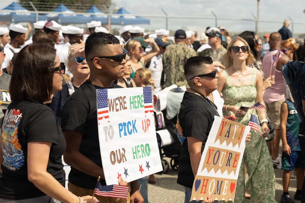 A military homecoming event where families and loved ones gather to welcome returning service members. Two boys in the foreground hold handmade signs reading 'We're Here to Pick Up Our Hero' and 'Yay! You're Home,' decorated with American flag motifs. The background shows a crowd of civilians and Navy personnel in white uniforms.