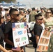 A military homecoming event where families and loved ones gather to welcome returning service members. Two boys in the foreground hold handmade signs reading 'We're Here to Pick Up Our Hero' and 'Yay! You're Home,' decorated with American flag motifs. The background shows a crowd of civilians and Navy personnel in white uniforms.