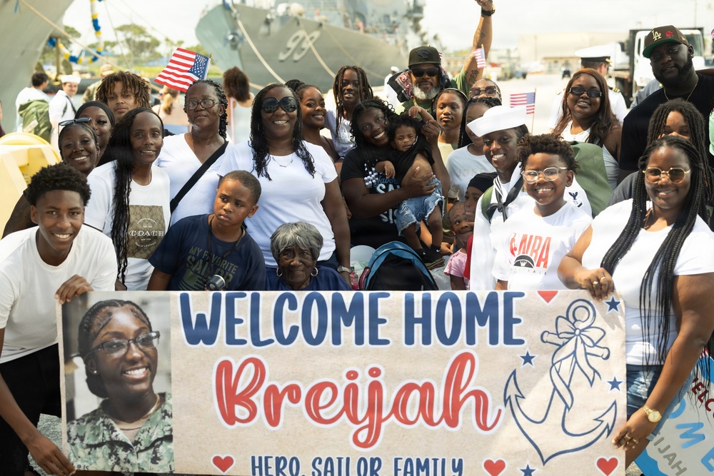 A large group of family and friends gathered at a naval pier to welcome home a sailor named Breijah. The group holds a sign reading 'Welcome Home Breijah - Hero, Sailor, Family' with an anchor graphic and a photo of the sailor. A naval vessel is visible in the background, and several people are waving small American flags.