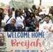 A large group of family and friends gathered at a naval pier to welcome home a sailor named Breijah. The group holds a sign reading 'Welcome Home Breijah - Hero, Sailor, Family' with an anchor graphic and a photo of the sailor. A naval vessel is visible in the background, and several people are waving small American flags.