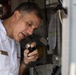 A senior naval officer in white dress uniform is speaking into a handheld microphone or radio handset, likely aboard a naval vessel. The officer displays gold shoulder boards indicating high rank, possibly an admiral or senior captain. He appears to be making an announcement or communicating via ship's intercom or radio system.
