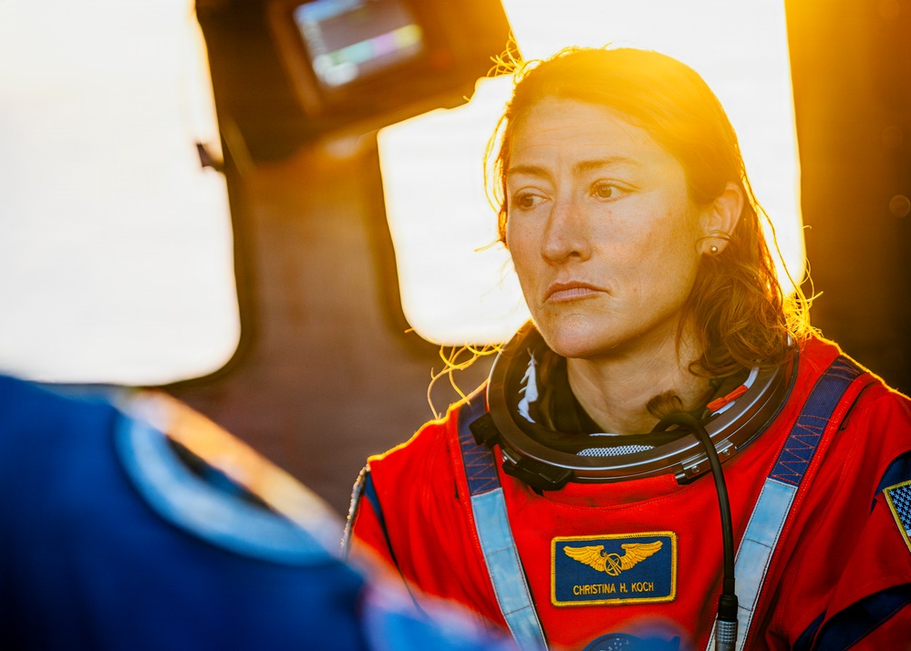 A person wearing a red flight suit with a name badge reading 'Christina H. Koch' and NASA astronaut wings insignia is visible in the foreground. The image is dramatically backlit with warm golden light, creating a cinematic atmosphere. A second person in a blue suit is partially visible in the foreground left.