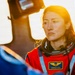 A person wearing a red flight suit with a name badge reading 'Christina H. Koch' and NASA astronaut wings insignia is visible in the foreground. The image is dramatically backlit with warm golden light, creating a cinematic atmosphere. A second person in a blue suit is partially visible in the foreground left.