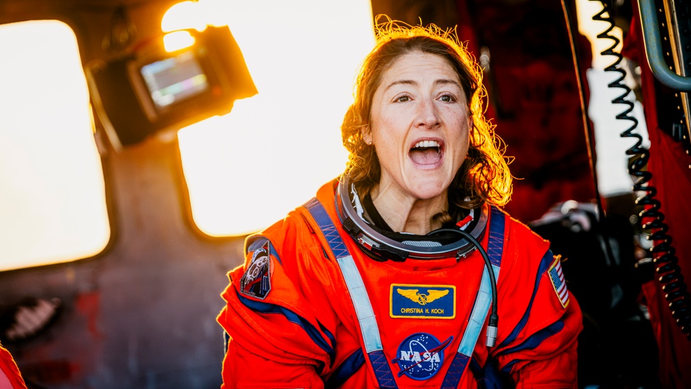 A NASA astronaut wearing an orange launch/entry suit is photographed with an expression of excitement or surprise. The name tag on the suit reads 'Christina H. Koch', identifying her as NASA astronaut Christina Koch. The image is dramatically lit with warm golden backlighting, creating a striking portrait.