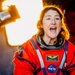 A NASA astronaut wearing an orange launch/entry suit is photographed with an expression of excitement or surprise. The name tag on the suit reads 'Christina H. Koch', identifying her as NASA astronaut Christina Koch. The image is dramatically lit with warm golden backlighting, creating a striking portrait.
