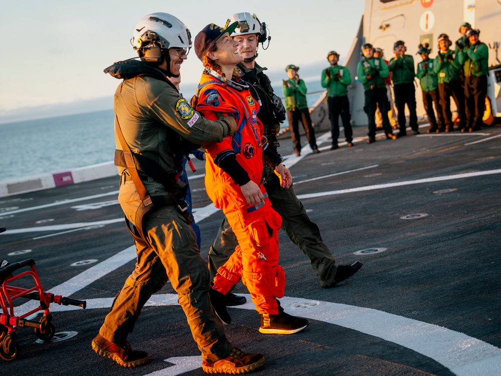 A person wearing an orange NASA flight suit is being assisted across what appears to be a naval vessel's flight deck by two personnel in military gear. A large group of crew members dressed in green uniforms are visible in the background, many appearing to applaud. The setting appears to be at sea during golden hour lighting conditions.