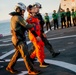 A person wearing an orange NASA flight suit is being assisted across what appears to be a naval vessel's flight deck by two personnel in military gear. A large group of crew members dressed in green uniforms are visible in the background, many appearing to applaud. The setting appears to be at sea during golden hour lighting conditions.