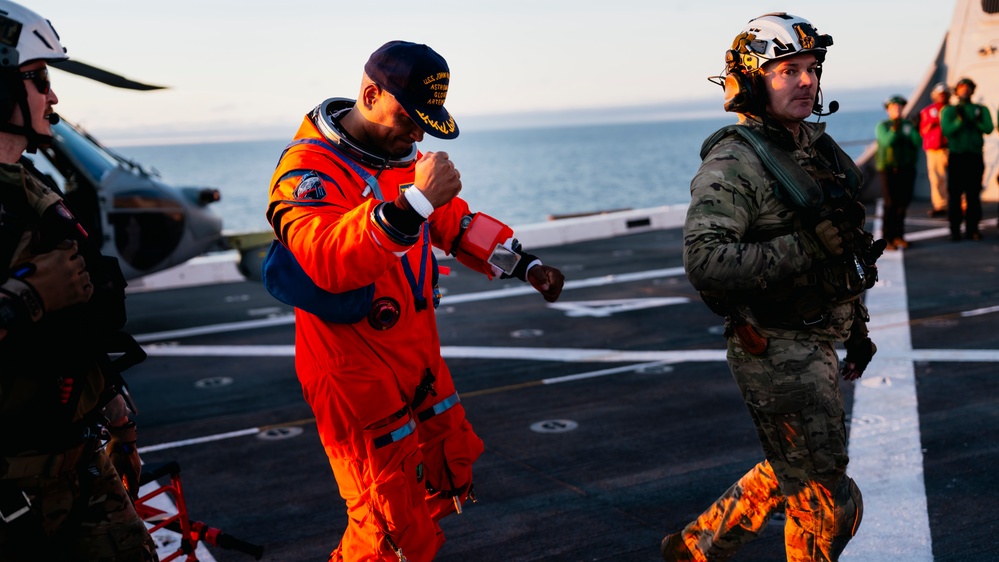 A person wearing an orange NASA-style astronaut flight suit and a navy cap is walking on what appears to be a naval vessel's flight deck, accompanied by military personnel in camouflage uniforms and tactical gear. The scene appears to be a post-splashdown recovery operation or training exercise, with the ocean visible in the background. The warm golden lighting suggests either sunrise or sunset conditions.
