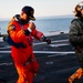 A person wearing an orange NASA-style astronaut flight suit and a navy cap is walking on what appears to be a naval vessel's flight deck, accompanied by military personnel in camouflage uniforms and tactical gear. The scene appears to be a post-splashdown recovery operation or training exercise, with the ocean visible in the background. The warm golden lighting suggests either sunrise or sunset conditions.