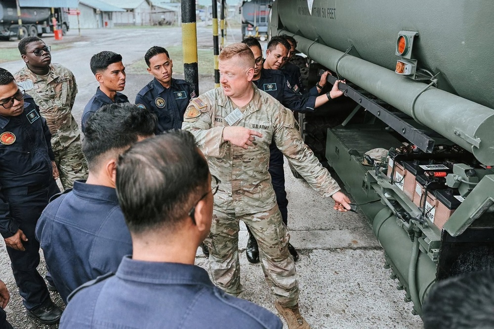 A U.S. Army soldier is conducting a training or briefing session for a group of foreign military or security personnel next to a large military vehicle, likely a fuel tanker. The soldier, whose name tape reads 'NEEL' and wears U.S. Army OCP (Operational Camouflage Pattern) uniform, is pointing at components on the side of the vehicle. The attendees are dressed in dark navy/black uniforms, suggesting they are from a partner nation's military or security force.