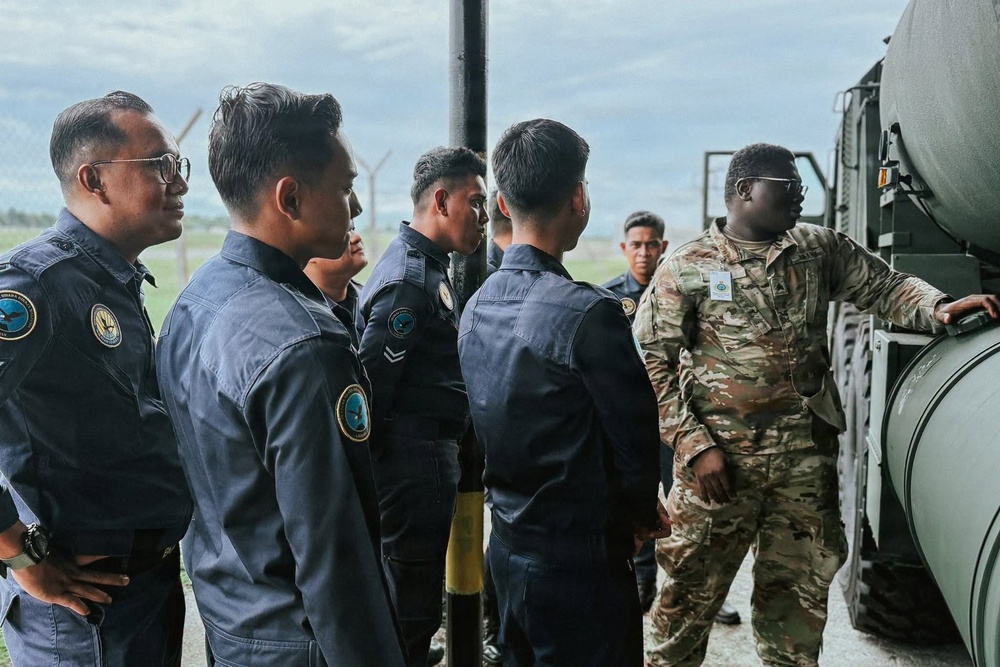A military training or demonstration session is taking place outdoors near what appears to be a military airfield or base. A soldier in MultiCam/OCP camouflage uniform is briefing or demonstrating equipment to a group of personnel wearing dark blue flight suits with unit patches. The interaction suggests an exchange program, joint exercise, or equipment familiarization training between different military forces.