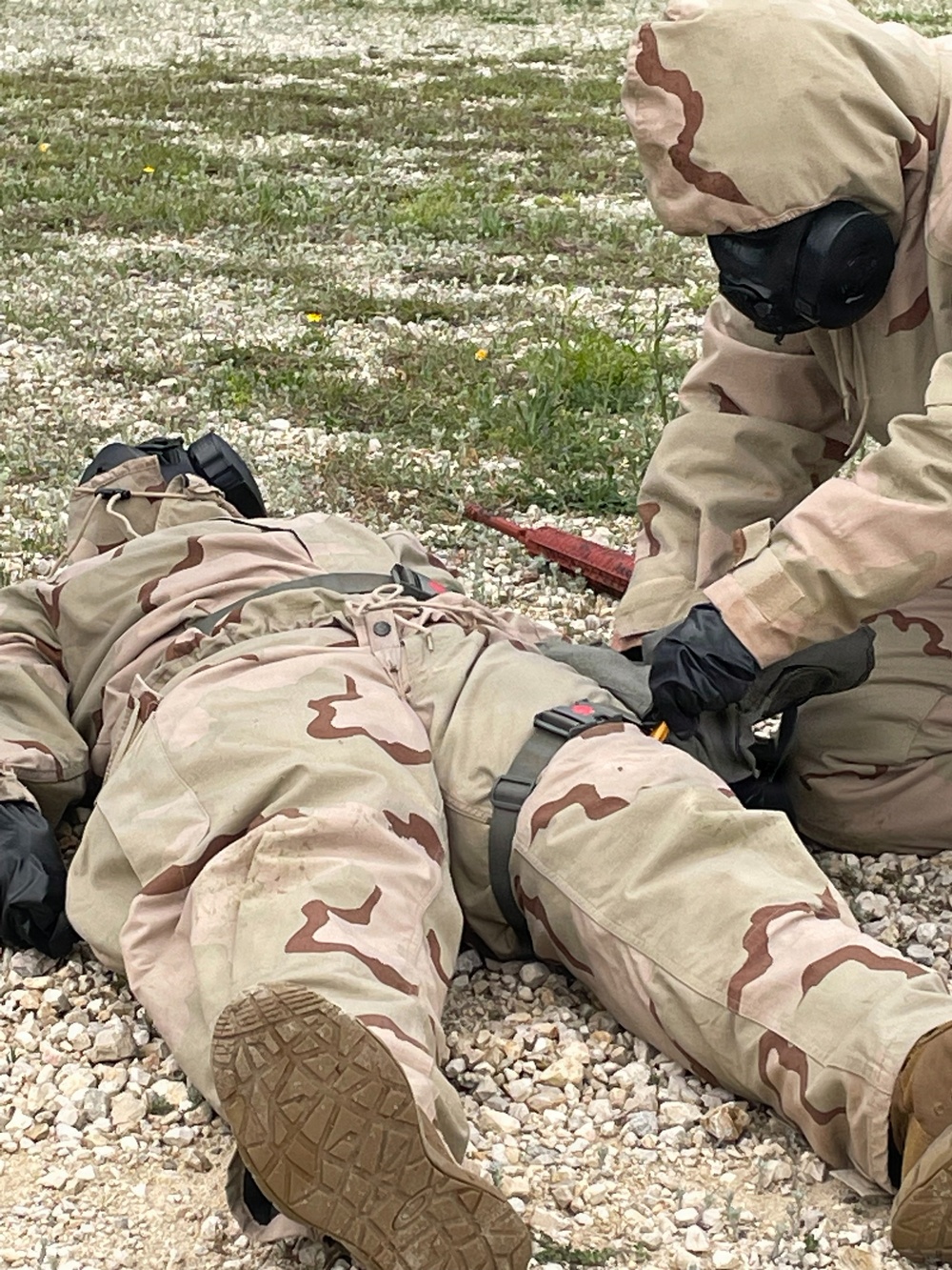 Two individuals wearing desert camouflage (DCU/three-color desert pattern) uniforms are shown on a gravel and sparse grass surface. One person is lying prone on the ground while another, wearing a full-face gas mask and black gloves, kneels over them appearing to administer aid or conduct a search. This appears to be a military training exercise, likely involving CBRN (Chemical, Biological, Radiological, Nuclear) casualty care procedures.