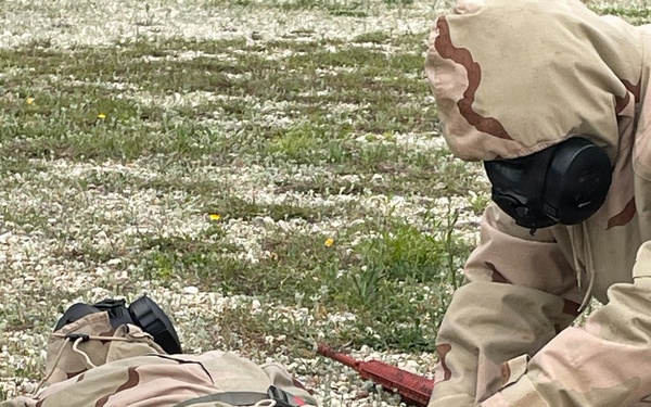 Two individuals wearing desert camouflage (DCU/three-color desert pattern) uniforms are shown on a gravel and sparse grass surface. One person is lying prone on the ground while another, wearing a full-face gas mask and black gloves, kneels over them appearing to administer aid or conduct a search. This appears to be a military training exercise, likely involving CBRN (Chemical, Biological, Radiological, Nuclear) casualty care procedures.