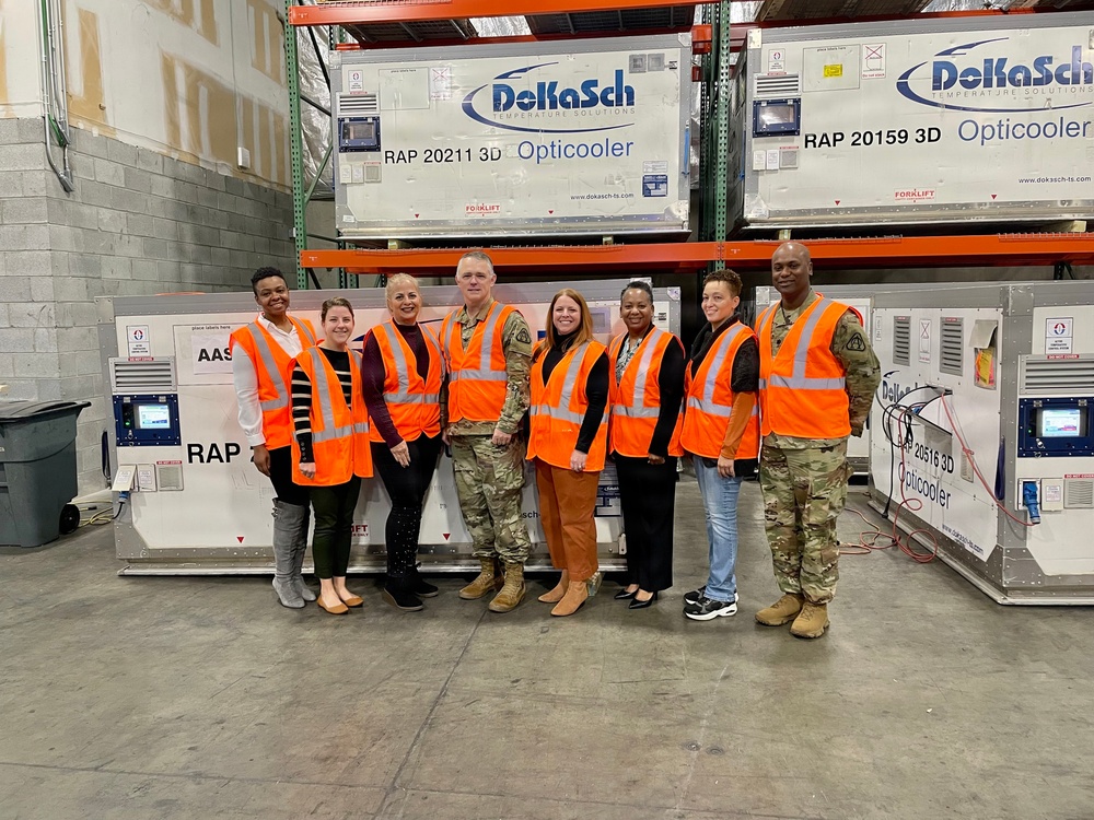 A group of eight people pose for a photo inside a warehouse facility in front of DoKaSch Temperature Solutions 'Opticooler' units. The group includes both military personnel in camouflage uniforms and civilians, all wearing orange high-visibility safety vests. The setting appears to be a logistics or cargo facility housing specialized temperature-controlled shipping containers.