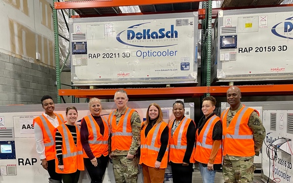 A group of eight people pose for a photo inside a warehouse facility in front of DoKaSch Temperature Solutions 'Opticooler' units. The group includes both military personnel in camouflage uniforms and civilians, all wearing orange high-visibility safety vests. The setting appears to be a logistics or cargo facility housing specialized temperature-controlled shipping containers.