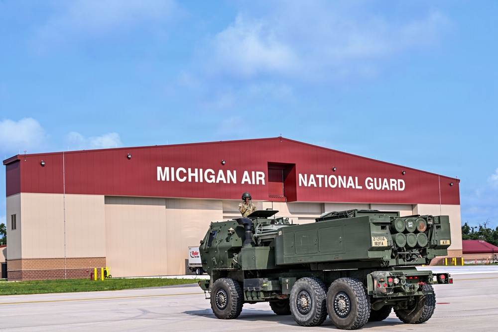 A military rocket artillery vehicle is positioned on a tarmac in front of a large red and tan hangar building marked 'Michigan Air National Guard.' A soldier in combat gear is visible operating or manning the vehicle. The image appears to be an official military documentation or public affairs photograph.