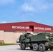 A military rocket artillery vehicle is positioned on a tarmac in front of a large red and tan hangar building marked 'Michigan Air National Guard.' A soldier in combat gear is visible operating or manning the vehicle. The image appears to be an official military documentation or public affairs photograph.