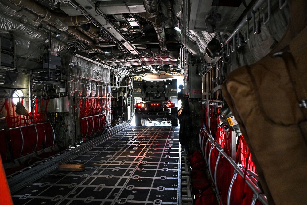 The image shows the interior cargo hold of a large military transport aircraft, likely a C-17 Globemaster III or C-5 Galaxy, viewed from front to rear. A military wheeled vehicle, possibly an HIMARS or similar rocket artillery system, is being loaded or unloaded through the open rear ramp. Red cargo restraint straps and tie-down rings line both sides of the cargo floor.