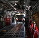 The image shows the interior cargo hold of a large military transport aircraft, likely a C-17 Globemaster III or C-5 Galaxy, viewed from front to rear. A military wheeled vehicle, possibly an HIMARS or similar rocket artillery system, is being loaded or unloaded through the open rear ramp. Red cargo restraint straps and tie-down rings line both sides of the cargo floor.