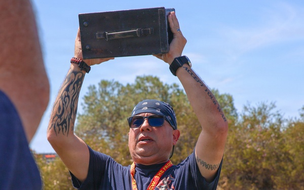 A middle-aged man with tattoos on both arms is holding a dark metal box (appears to be an ammunition can) overhead with both hands, likely performing a fitness exercise. He is wearing a navy blue t-shirt, bandana, sunglasses, a watch, and a lanyard with an ID badge. The setting appears to be an outdoor athletic track or sports field on a sunny day.