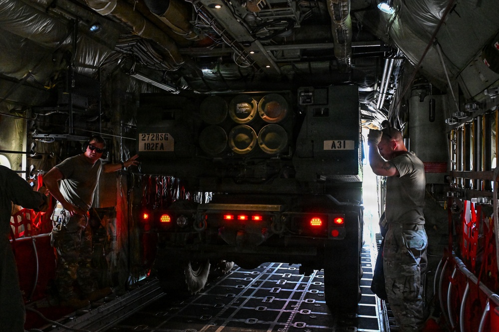 Military personnel are loading or unloading a rocket artillery system, identified by markings as belonging to 272RSG/182FA, inside the cargo hold of a large military transport aircraft. The vehicle appears to be an M270 Multiple Launch Rocket System (MLRS) or similar rocket artillery platform, visible by its distinctive launcher pod configuration. Two soldiers in military uniforms are guiding the vehicle within the aircraft's cargo bay.