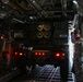 Military personnel are loading or unloading a rocket artillery system, identified by markings as belonging to 272RSG/182FA, inside the cargo hold of a large military transport aircraft. The vehicle appears to be an M270 Multiple Launch Rocket System (MLRS) or similar rocket artillery platform, visible by its distinctive launcher pod configuration. Two soldiers in military uniforms are guiding the vehicle within the aircraft's cargo bay.