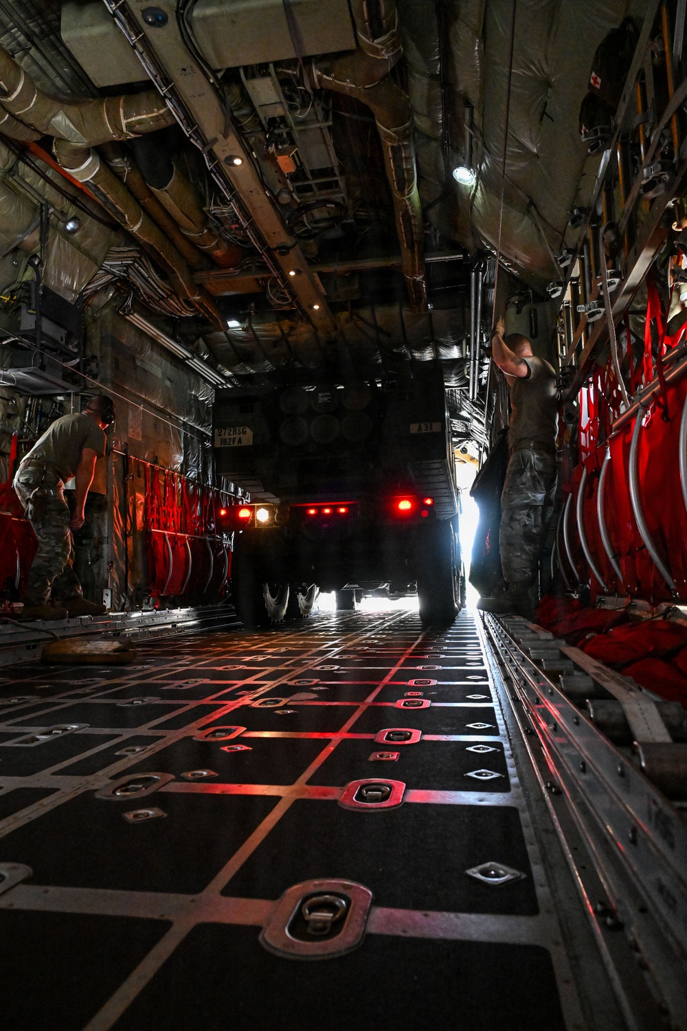 The interior of a large military transport aircraft, likely a C-17 Globemaster or C-130 Hercules, showing a military vehicle (HMMWV/Humvee) being loaded or unloaded via the rear ramp. The cargo bay is illuminated by red ambient lighting and the vehicle's brake lights, with two personnel in military uniforms guiding the operation. The ramp and floor feature standard military cargo tie-down rings and grid markings.