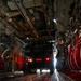 The interior of a large military transport aircraft, likely a C-17 Globemaster or C-130 Hercules, showing a military vehicle (HMMWV/Humvee) being loaded or unloaded via the rear ramp. The cargo bay is illuminated by red ambient lighting and the vehicle's brake lights, with two personnel in military uniforms guiding the operation. The ramp and floor feature standard military cargo tie-down rings and grid markings.