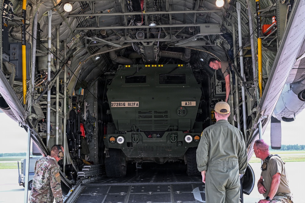 A large military cargo aircraft, likely a C-17 Globemaster or C-130, is being loaded with a military wheeled vehicle marked '272RSG 182FA' and 'A31'. Several personnel in military and flight suits are overseeing the loading operation from the rear cargo ramp. The scene appears to take place on an active airfield tarmac.