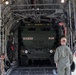 A large military cargo aircraft, likely a C-17 Globemaster or C-130, is being loaded with a military wheeled vehicle marked '272RSG 182FA' and 'A31'. Several personnel in military and flight suits are overseeing the loading operation from the rear cargo ramp. The scene appears to take place on an active airfield tarmac.