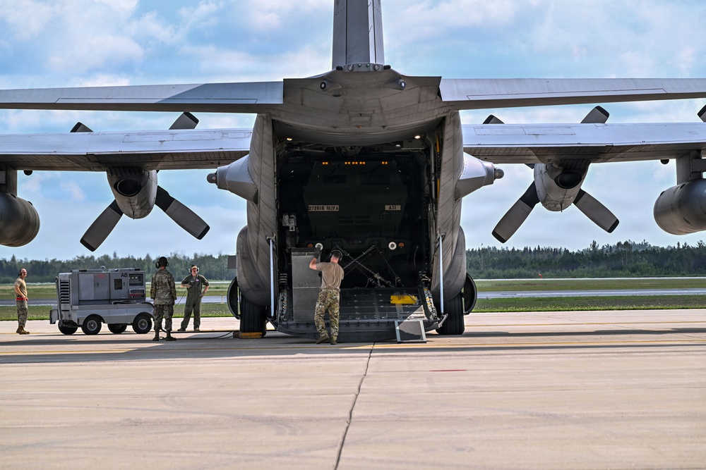 A large military transport aircraft, consistent with a C-130 Hercules, is parked on a tarmac with its rear cargo ramp lowered. Several military personnel in camouflage uniforms are visible near the aircraft, with one individual standing on the ramp appearing to direct or signal, while others stand near ground support equipment. The setting appears to be a military airfield surrounded by trees under partly cloudy skies.
