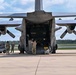 A large military transport aircraft, consistent with a C-130 Hercules, is parked on a tarmac with its rear cargo ramp lowered. Several military personnel in camouflage uniforms are visible near the aircraft, with one individual standing on the ramp appearing to direct or signal, while others stand near ground support equipment. The setting appears to be a military airfield surrounded by trees under partly cloudy skies.