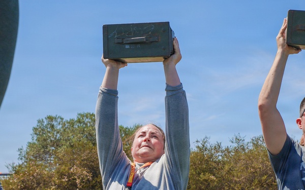 A group of people are participating in an outdoor physical fitness exercise on a sports field, lifting military-style ammunition cans overhead. The central figure is a woman wearing a gray long-sleeve shirt and a lanyard with a badge, straining to hold the metal box above her head. A man to her right in a gray t-shirt is also lifting a similar ammo can, suggesting a group fitness or military-style challenge event.