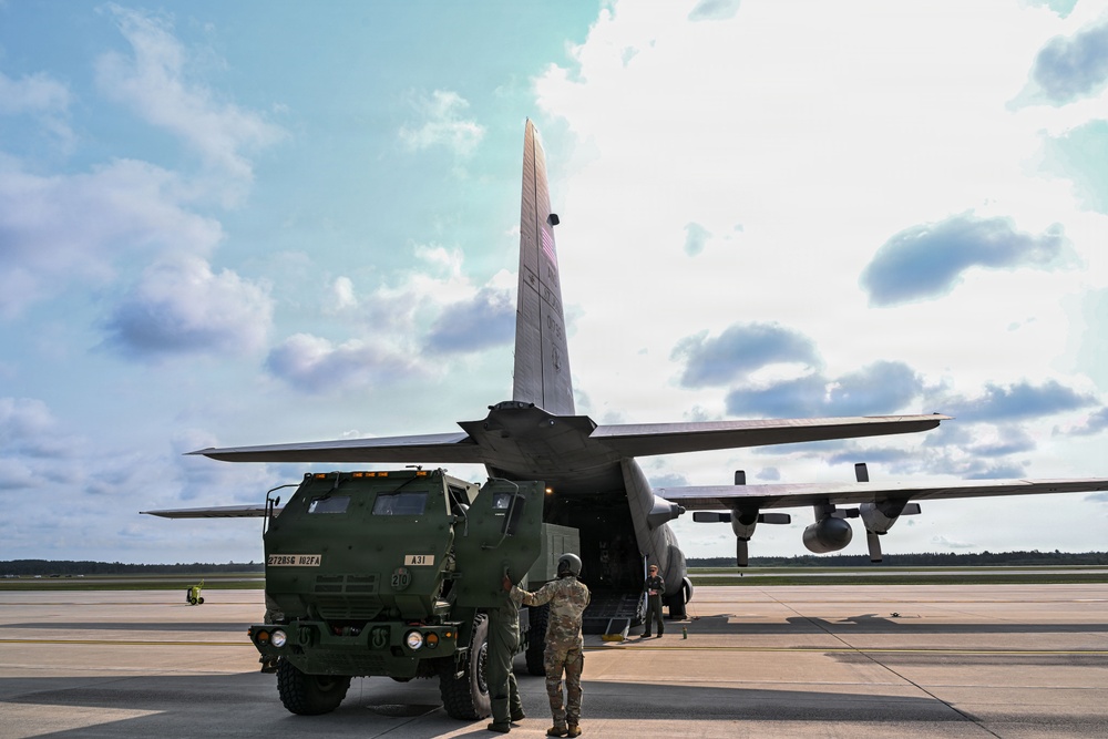 A military tactical vehicle is being loaded onto a large military transport aircraft on an airfield tarmac. Personnel in military uniforms are directing the vehicle as it backs or drives into the rear cargo ramp of what appears to be a C-130 Hercules aircraft. The scene depicts a standard military airlift or deployment operation under partly cloudy skies.