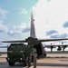 A military tactical vehicle is being loaded onto a large military transport aircraft on an airfield tarmac. Personnel in military uniforms are directing the vehicle as it backs or drives into the rear cargo ramp of what appears to be a C-130 Hercules aircraft. The scene depicts a standard military airlift or deployment operation under partly cloudy skies.