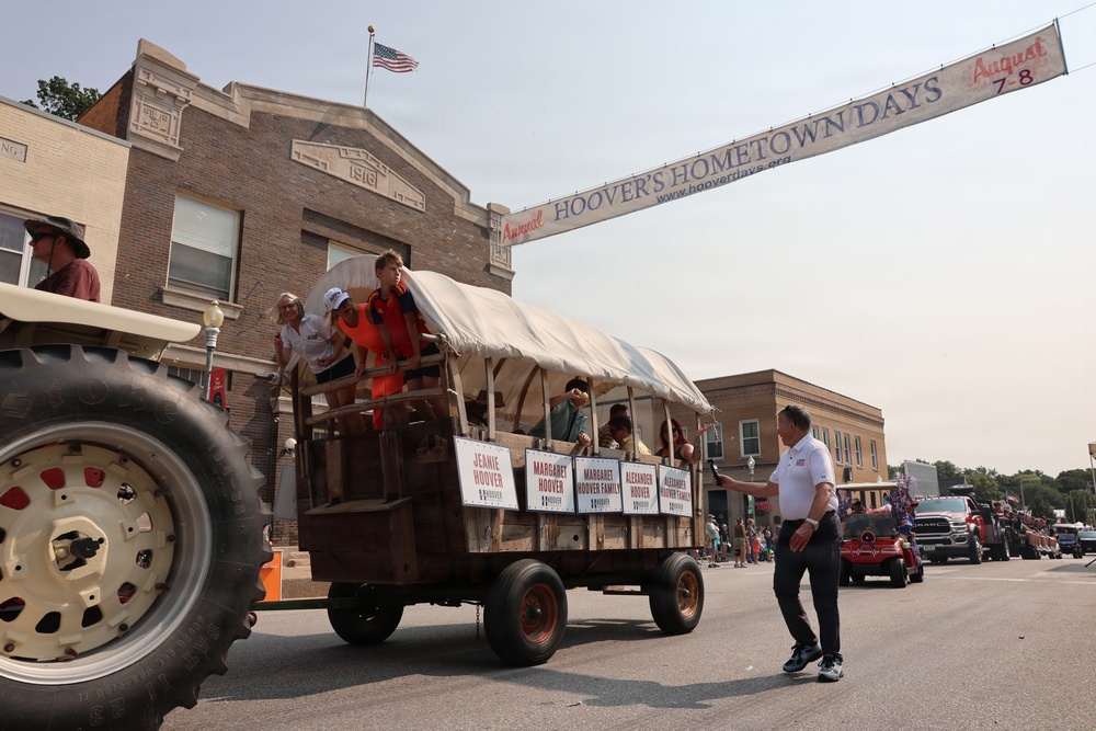 A parade is taking place during 'Hoover's Hometown Days' annual festival, held August 7-8, as indicated by a banner spanning the street. A covered wagon-style float pulled by a tractor carries several people, with signs on the side reading 'Jeanie Hoover,' 'Margaret Hoover,' 'Alexander Hoover,' and 'Alexander Hoover Family.' The event appears to be held in a small American town with historic brick buildings, an American flag, and crowds lining the street.