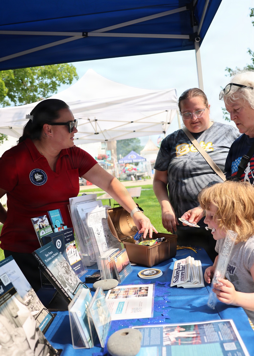 A representative wearing a red polo shirt with a Naval History and Heritage Command patch is staffing an outdoor information booth at what appears to be a public event or fair. She is engaging with visitors including two adults and a child, showing items from a decorative chest on a blue-draped table covered with Navy-related brochures and materials. A sign reading 'Sailors have more fun!' is visible among the display items.
