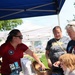 A representative wearing a red polo shirt with a Naval History and Heritage Command patch is staffing an outdoor information booth at what appears to be a public event or fair. She is engaging with visitors including two adults and a child, showing items from a decorative chest on a blue-draped table covered with Navy-related brochures and materials. A sign reading 'Sailors have more fun!' is visible among the display items.