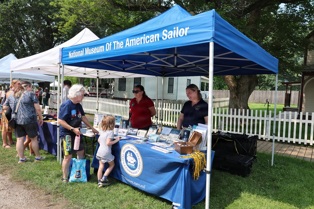 An outdoor event booth for the National Museum of The American Sailor, operated under the Naval History and Heritage Command (www.history.navy.mil). Two staff members in uniform polo shirts are staffing a blue-draped table with informational materials and brochures. Visitors, including an elderly woman and a small child, are interacting with the booth staff in what appears to be a community or heritage fair setting.