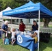 An outdoor event booth for the National Museum of The American Sailor, operated under the Naval History and Heritage Command (www.history.navy.mil). Two staff members in uniform polo shirts are staffing a blue-draped table with informational materials and brochures. Visitors, including an elderly woman and a small child, are interacting with the booth staff in what appears to be a community or heritage fair setting.