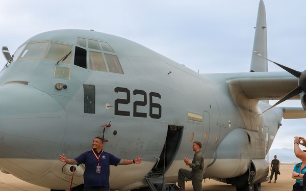 A military transport aircraft marked '226' is displayed on a tarmac, likely during an air show or public open house event. Civilians are posing for photographs in front of the aircraft while a uniformed crew member stands near the open side door. Multiple personnel and additional aircraft are visible in the background.