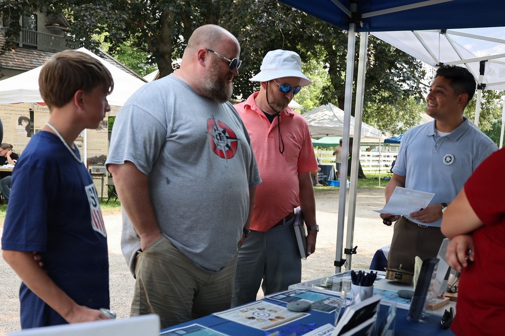 An outdoor event or fair where visitors are gathered around an informational booth or display table under a blue canopy tent. A representative holding papers appears to be engaging with three visitors, including a young boy and two adult men. The setting includes multiple vendor or exhibit tents visible in the background among trees.