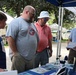An outdoor event or fair where visitors are gathered around an informational booth or display table under a blue canopy tent. A representative holding papers appears to be engaging with three visitors, including a young boy and two adult men. The setting includes multiple vendor or exhibit tents visible in the background among trees.