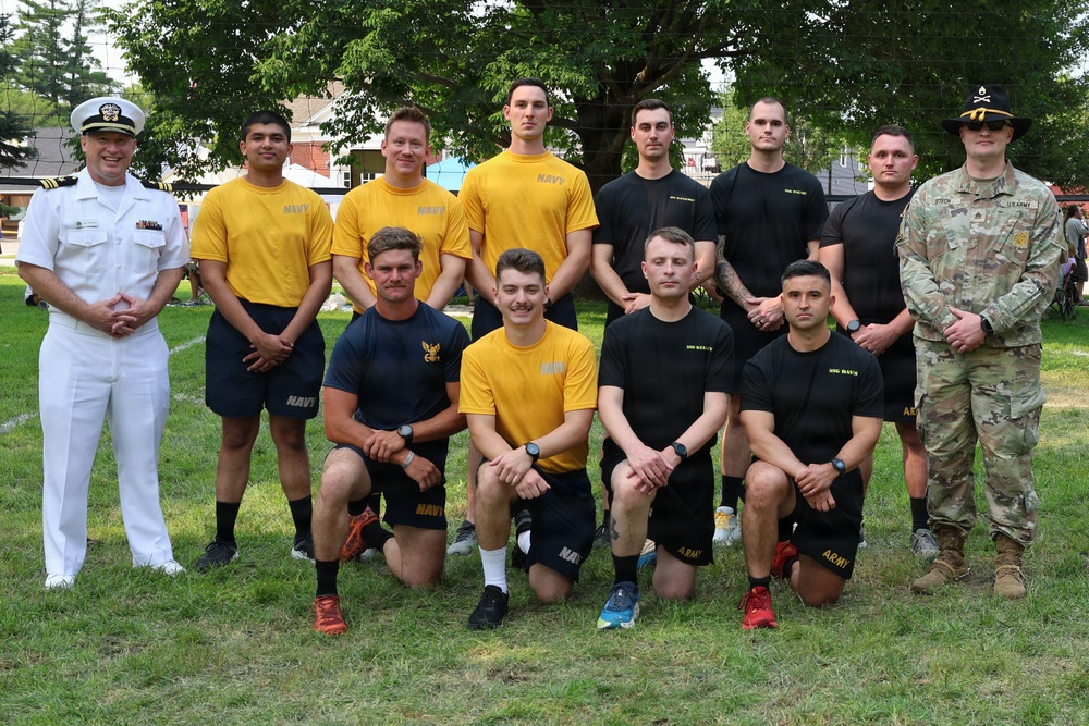 A group of approximately 11 military personnel pose for a formal group photo on a grass field. The group includes members from both the U.S. Navy (wearing gold/yellow 'NAVY' shirts and navy blue shorts) and U.S. Army (wearing black shirts and camouflage uniform). A senior Naval officer in dress whites stands on the far left, and an Army soldier in full ACU/OCP camouflage with a distinctive hat stands on the far right.